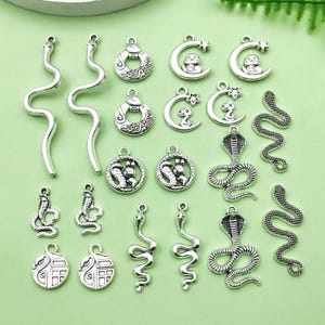 Pode incluir: Uma coleção de pingentes de metal prateados com desenhos de cobra e lua crescente. Os pingentes são perfeitos para fazer joias, scrapbooking e outros projetos de artesanato.