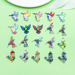 May include: A collection of twenty metal charms shaped like hummingbirds and small birds, featuring colorful enamel designs with various patterns, stripes, and floral motifs. Each charm includes small loops at the top and bottom for attachment.