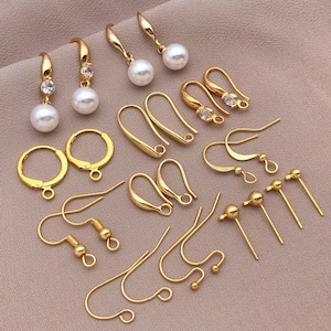 Op de afbeelding: Een verzameling goudkleurige oorbelcomponenten, waaronder haken, ringen en pinnen. Sommige oorbellen hebben heldere edelstenen en witte imitatieparels. De componenten zijn gerangschikt op een roze-beige stoffen oppervlak.