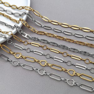 Könnte beinhalten: Eine Auswahl an gold- und silberfarbenen Kettenhalsketten. Die Ketten weisen verschiedene Gliederdesigns auf, darunter ovale, rechteckige und herzförmige Glieder. Die Halsketten werden auf einer hellgrauen Oberfläche präsentiert, wobei einige Ketten teilweise auf einem weißen Teller liegen.