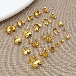 May include: A collection of various gold-colored jewelry findings, including crimp beads, end caps, and clasps. The components are arranged on a neutral-toned surface, showcasing their different shapes and sizes. These findings are used for jewelry making.