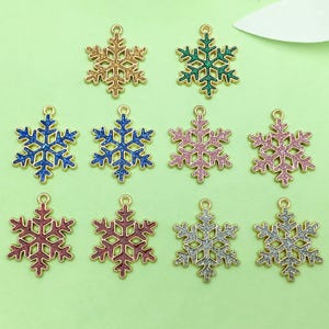 Op de afbeelding: Een verzameling sneeuwvlok-bedels in verschillende kleuren, waaronder goud, groen, blauw, roze, rood en zilver. Elke bedel heeft een goudkleurige omtrek en een lus om te bevestigen. De sneeuwvlokken zijn glinsterend.