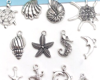 May include: A collection of 14 silver-toned metal charms featuring various sea life and shell designs. The charms include a turtle, seashells, starfish, a seahorse, a dolphin, and a sun.