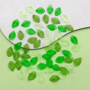 May include: A collection of green and clear plastic leaf-shaped charms with a hole at the top. The charms are scattered on a white and green surface.