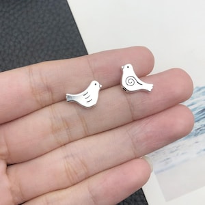 Könnte beinhalten: Zwei silberne Vogel-Anhänger mit schlichtem Design. Die Vögel sind in entgegengesetzte Richtungen gerichtet und haben eine leicht erhabene Oberfläche.