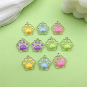 Op de afbeelding: Tien kleurrijke pootjescharms met een transparante achtergrond. De charms zijn in verschillende kleuren, waaronder geel, blauw, roze, paars en groen.