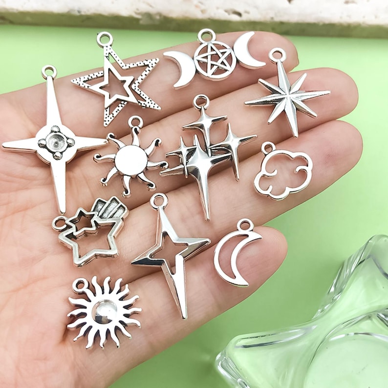 K&ouml;nnte beinhalten: Eine Sammlung silberfarbener Anh&auml;nger in verschiedenen Himmelsformen. Die Anh&auml;nger umfassen Sterne, Monde, Sonnen und Wolken. Jeder Anh&auml;nger hat eine kleine Schlaufe zum Befestigen an Schmuck oder anderen Bastelarbeiten. Die Anh&auml;nger werden vor einem hellgr&uuml;nen Hintergrund pr&auml;sentiert.