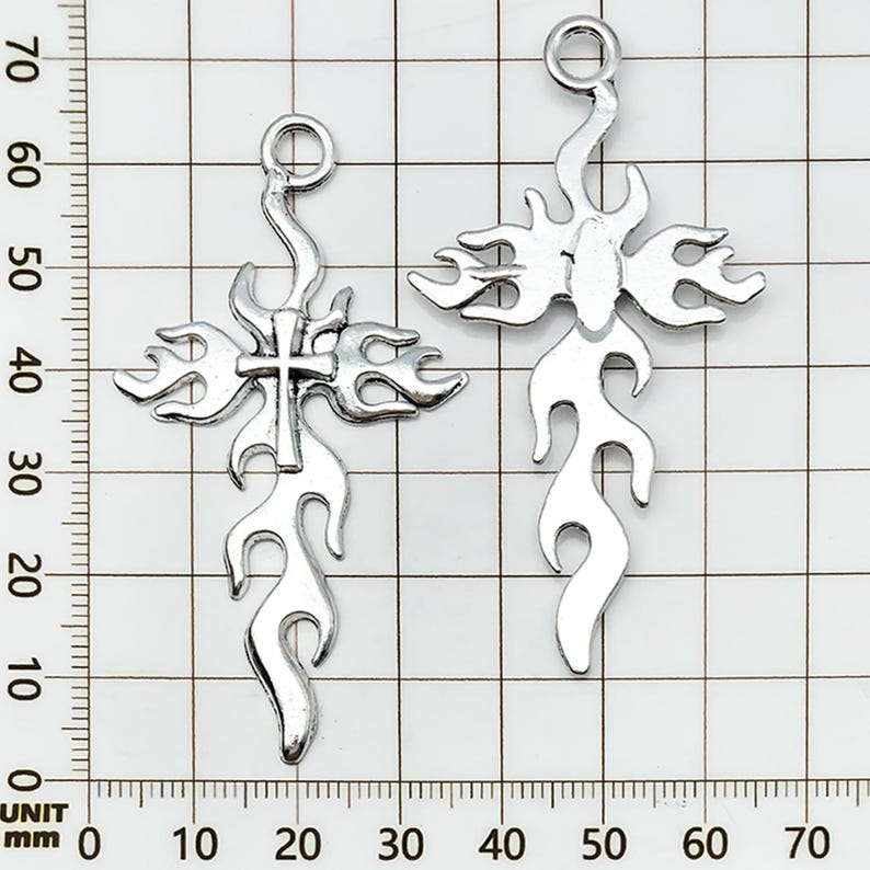 May include: Two silver-colored cross pendants with a flame design. Each cross has a loop at the top for hanging and measures approximately 2.5 inches in length. The crosses are displayed against a grid background.