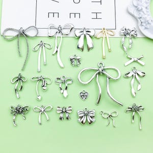 Könnte beinhalten: Eine Sammlung von silberfarbenen Schleifenanhängern in verschiedenen Designs, darunter klassische Schleifen, Bänder und herzförmige Akzente. Die Anhänger sind auf einer hellgrünen Oberfläche angeordnet, ideal für Schmuckherstellung oder Bastelprojekte.