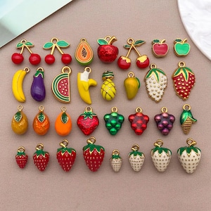 Puede incluir: Colección de colgantes con forma de frutas coloridas, incluyendo cerezas, manzanas, fresas, plátanos y uvas. Cada colgante tiene un lazo dorado para sujetarlo a joyas u otras manualidades. Los colgantes están pintados con colores brillantes y diseños detallados.