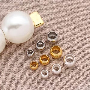 May include: An assortment of jewelry-making beads in gold and silver tones. The beads are round and cylindrical, with varying sizes. A large white pearl and gold clasp are also visible, suggesting a jewelry-making theme.