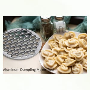 May include: A silver aluminum dumpling maker tool with a honeycomb design sits next to a plate of cooked dumplings. The text "Aluminum Dumpling Maker Tool" is visible below the tool. Two glass shakers are in the background.