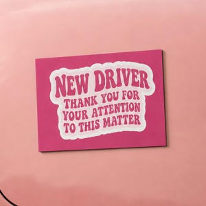 May include: A rectangular sticker with a pink background and white wavy-edged text that reads NEW DRIVER THANK YOU FOR YOUR ATTENTION TO THIS MATTER, attached to the trunk of a light pink car with a chrome bumper and a red taillight.