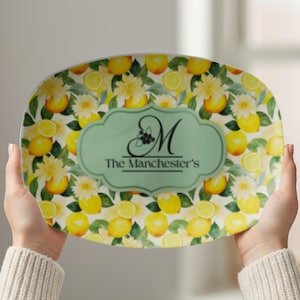 May include: An oval serving platter with a lemon and floral design. The platter features a light green central emblem with the letter "M" and the text "The Manchester's". Yellow lemons and white flowers are scattered across the surface.