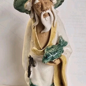 Op de afbeelding: Een keramieken beeldje van een oude man in een witte jurk en een groene hoed, met een groene vis in zijn hand. Het beeldje heeft een lange witte baard en staat op een donkere basis. De jurk heeft gele accenten.
