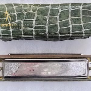 May include: A Hohner Chromonica harmonica with a silver finish and decorative scrollwork, resting below a green, textured case. The case has a reptile-like pattern and the text "Hohner-Chromonica" is visible.