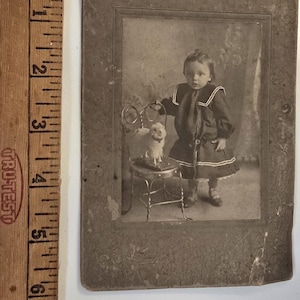 Puede incluir: Fotografía vintage en tonos sepia de un niño pequeño con un vestido de marinero, de pie junto a una pequeña silla con un animal de peluche. El niño lleva un vestido oscuro con ribetes blancos y un cuello de marinero a juego. La fotografía está en un marco rectangular.