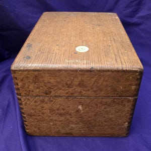 May include: A rectangular wooden box with visible finger joints at the corners and a natural wood grain finish. The top surface features a small circular sticker with the number 65 and the text Shaw-Walker embossed in gold lettering on the lid.