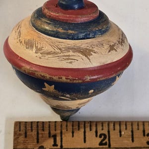 Puede incluir: Una peonza de madera vintage, pintada en rojo, blanco y azul. La parte superior tiene una forma redondeada con una base puntiaguda y mide aproximadamente 6,35 cm de alto. El diseño incluye estrellas y rayas, evocando un tema patriótico.