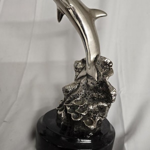 May include: A silver-colored dolphin sculpture leaping from a textured rock formation, mounted on a black, circular base. The dolphin is smooth and polished, with a streamlined body. The sculpture is a decorative item.