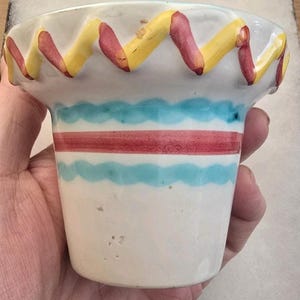 May include: A small, white ceramic container with a wavy yellow and red rim. The container has a red stripe and a blue wavy stripe around the middle. The container is held in a hand.