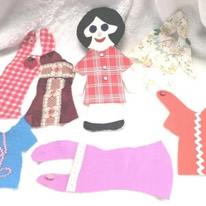 Könnte beinhalten: Eine Sammlung handgefertigter Puppenkleidung aus Stoff und eine Puppe. Die Puppe hat einen schwarzen ovalen Kopf, schwarze Haare und ein rot-weiß kariertes Kleid. Die Kleidung ist in verschiedenen Farben und Mustern gehalten.