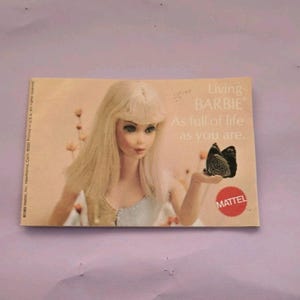 May include: A vintage Barbie advertisement featuring a doll with long blonde hair and a light blue dress. The text reads "Living BARBIE As full of life as you are." The Mattel logo is visible.