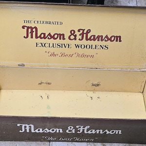 May include: An open, vintage rectangular box with a brown exterior and a light yellow interior. The box features the text "Mason & Hanson" in a decorative font, along with "Exclusive Woolens" and "The Best Woven". The box appears aged.