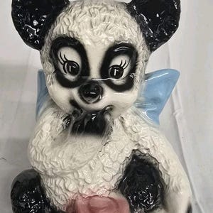 Puede incluir: Figura de cerámica de un oso panda con orejas, ojos y manchas negras, y cuerpo blanco. El panda sostiene un tarro de miel rosa con la palabra "HONEY". Una cinta azul claro está detrás de la cabeza del panda.