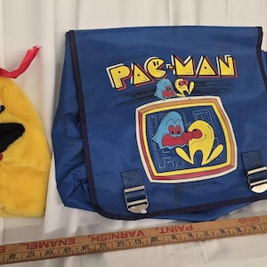 May include: A blue Pac-Man messenger bag featuring a Pac-Man graphic and a yellow plush character with a red ribbon. The bag has silver buckles and a ruler is at the bottom.