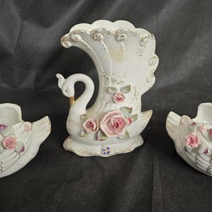May include: A set of three white ceramic swan ornaments. Two are open-topped, and one is a vase. Each features pink roses and gold detailing. The swan ornaments are decorative and suitable for home decor.