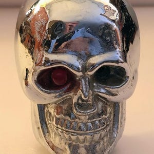 May include: A reflective chrome-finished skull ornament featuring a single embedded red circular stone in the right eye socket. The skull has a smooth, metallic surface with visible contours for the nose, teeth, and brow, creating a high-contrast look.