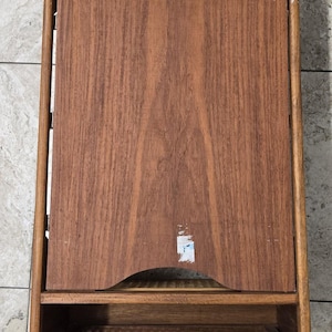 May include: A vintage wooden side table with a rectangular design. The table features a large, solid wood panel in the center, framed by a lighter wood structure. It has a woven cane shelf and a similar top shelf. The wood grain is visible.