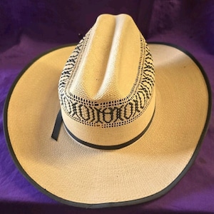 Pode incluir: Um chapéu de cowboy de palha com coroa e aba bege claro. O chapéu apresenta uma faixa preta estampada ao redor da coroa e uma borda preta ao longo da borda. Uma fita preta é presa na lateral.