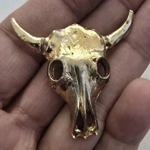 May include: A metallic gold-colored bull skull pendant featuring a textured surface, prominent horns, and hollow eye sockets. The object has a weathered, cast metal appearance with visible indentations and a reflective finish across the entire skull.