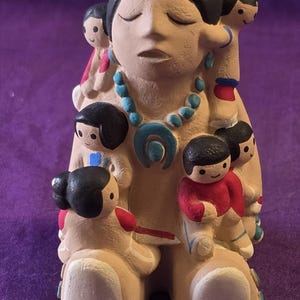 May include: A ceramic figurine depicting a seated figure surrounded by several smaller figures. The central figure wears a turquoise necklace and has closed eyes. The smaller figures are in various poses, some on the central figure's shoulders. The figurine is painted in earth tones with accents of red and blue.