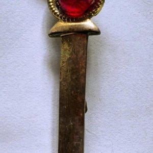 May include: A sword-shaped brooch featuring a gold-toned hilt with a textured grip and a crossguard. A large, round, faceted red gemstone is set in a gold-toned circular frame at the base of the hilt. The blade is a flat, dark, metallic brown.
