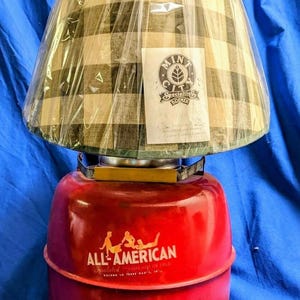 Könnte beinhalten: Eine rot-gelbe Lampe mit kariertem Lampenschirm. Der Lampensockel ist rot mit den Worten "ALL-AMERICAN" und "Insulated" bedruckt. Der Lampenschirm ist mit einer transparenten Plastikfolie mit einem Etikett mit der Aufschrift "Mint City Specialties" bedeckt.