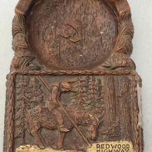Puede incluir: Placa de madera tallada en color marrón que presenta un relieve de una persona a caballo en un bosque, con el texto "Trees of Mystery California" y "Redwood Highway". La placa tiene un borde decorativo y una parte superior circular.