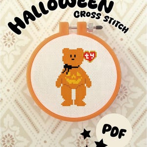 Può includere: Un motivo a punto croce in un telaio da ricamo arancione raffigurante un orsacchiotto arancione con un fiocco nero, un disegno di zucca sul petto e un'etichetta rossa a forma di cuore con le lettere ty. Il testo HALLOWEEN CROSS STITCH e PDF è presente.