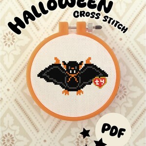 Può includere: Uno schema a punto croce in un telaio da ricamo arancione raffigurante un pipistrello nero in pixel art con orecchie, piedi e colletto arancioni, che tiene un'etichetta rossa a forma di cuore con le lettere ty. Il testo HALLOWEEN CROSS STITCH e PDF è visibile.