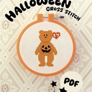 May include: A cross stitch pattern in an orange embroidery hoop featuring a brown bear with a black jack-o'-lantern face on its chest and a red heart-shaped tag with the letters ty. The text HALLOWEEN CROSS STITCH and PDF are visible.