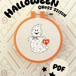 May include: A round orange embroidery hoop containing white Aida fabric with a cross-stitch pattern of a white ghost holding a red heart-shaped tag with the letters ty. The text HALLOWEEN CROSS STITCH and PDF are visible on the image.
