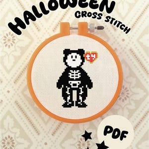 May include: An orange embroidery hoop containing white Aida fabric with a cross-stitch pattern of a black bear wearing a white skeleton costume and a red heart-shaped tag. The text HALLOWEEN CROSS STITCH and PDF are visible.