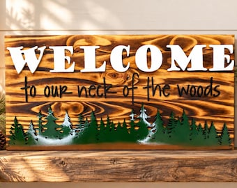 Puede incluir: Un letrero rectangular de madera con acabado quemado que presenta el texto 3D blanco WELCOME y el texto en cursiva negro to our neck of the woods. La sección inferior muestra una silueta de bosque verde superpuesta con detalles blancos.