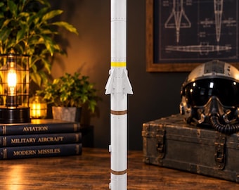 Puede incluir: Un modelo vertical de misil AIM-120A AMRAAM con cuerpo cilíndrico blanco, punta cónica, cuatro aletas centrales y cuatro aletas traseras. El misil está montado sobre una base cuadrada negra con una etiqueta que dice AIM-120A AMRAAM.