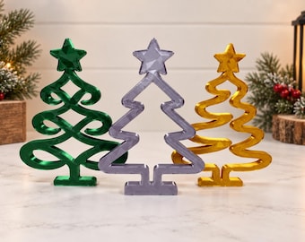 Puede incluir: Tres decoraciones de árbol de Navidad de pie en verde, morado claro y dorado. Cada árbol presenta un diseño estilizado de marco abierto con una estrella en la parte superior y una base rectangular. Las superficies tienen un acabado metálico liso y reflectante.