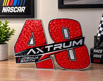 Puede incluir: Un letrero decorativo rojo y plateado con forma de número 48 y un patrón geométrico. El letrero presenta el texto AXTRUM en el centro y JOSHUA SMITH en un recuadro más pequeño en la parte inferior izquierda, todo con un borde plateado metálico.