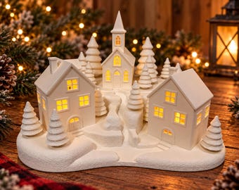 Puede incluir: Un pueblo navideño de cerámica blanca con dos casas y una iglesia central, iluminado desde el interior por luces amarillas cálidas. La escena incluye varios árboles de hoja perenne esculpidos y una base texturizada cubierta de nieve.