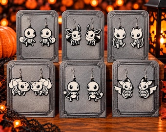 Puede incluir: Seis pares de pendientes colgantes con criaturas de dibujos animados con temática de esqueleto en blanco y negro. Cada par se muestra en una tarjeta rectangular gris con un borde decorativo y diseños de personajes con cajas torácicas.
